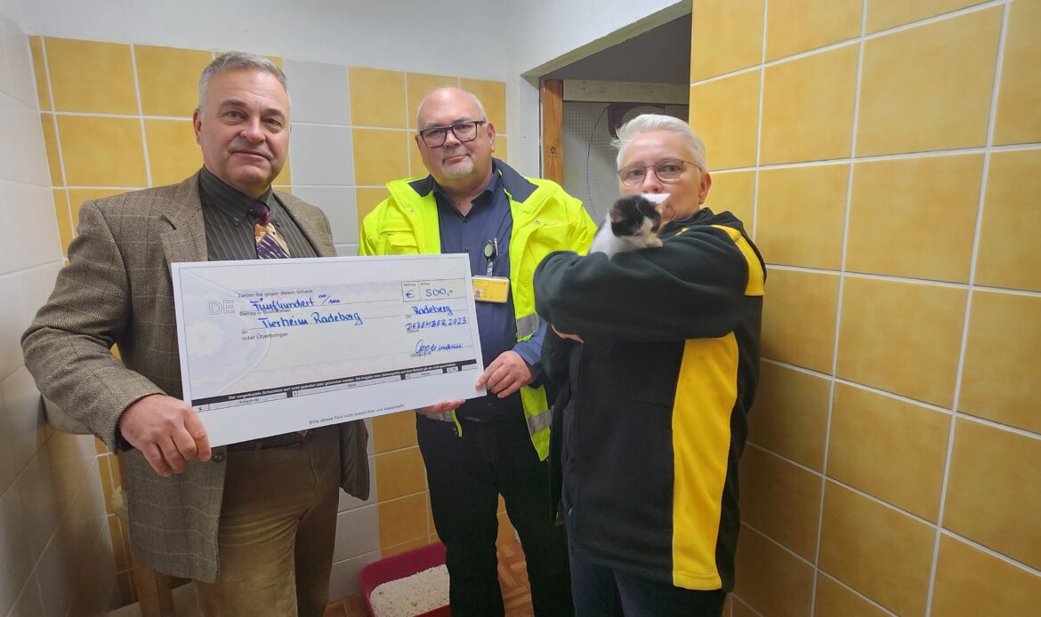 Drei Personen stehen in einem Raum des Tierheims, halten einen großen Spendenscheck und Sachspenden; eine Person trägt eine Katze auf dem Arm.