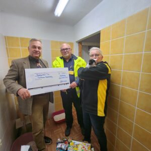 Drei Personen stehen in einem Raum des Tierheims, halten einen großen Spendenscheck und Sachspenden; eine Person trägt eine Katze auf dem Arm.